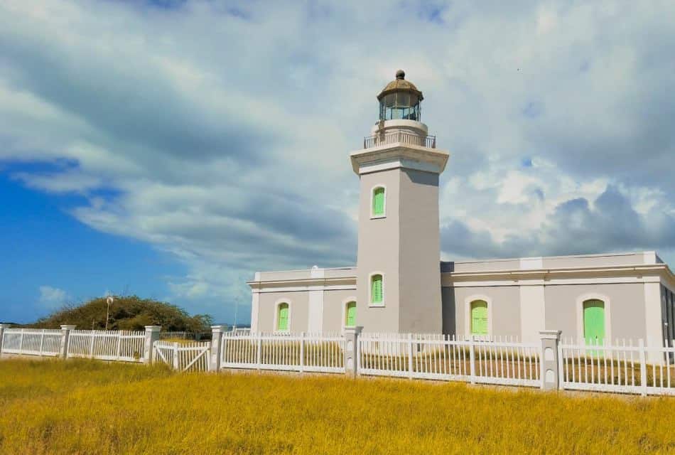Gray lighthouse with bright green windows surrounded by white fencing and yellow grass