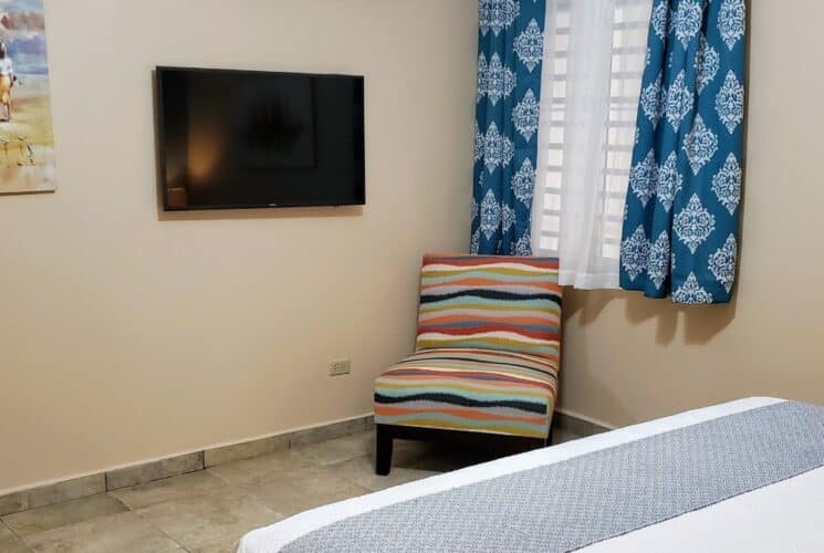 Bedroom with tan walls, tile flooring, white bedding, multicolored chair, and flatscreen TV