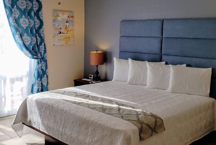 Bedroom with light blue and tan walls, tile flooring, blue upholstered headboard, white bedding, and large double door entrance with blue and white curtains