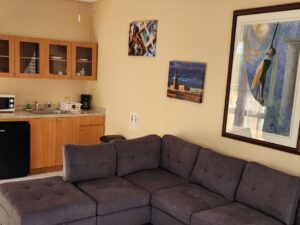 Large suite with light tan walls, tile flooring, upholstered gray sectional sofa, small kitchen area with wooden cabinets, mini fridge, microwave, sink, two queen beds and view into bathroom