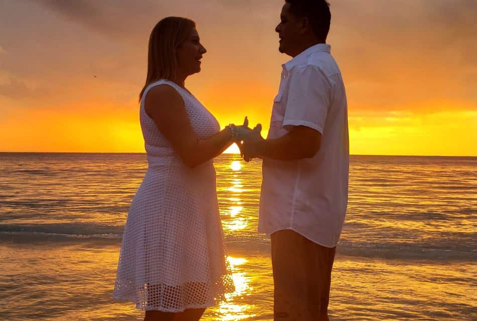 Woman in white dress and man wearing a white shirt holding hands and looking at each other with the ocean and setting sun in background