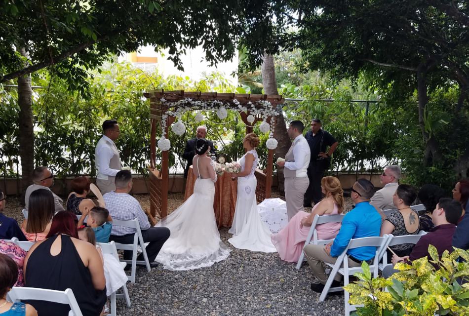 Two women wearing white dresses standing at a wooden arbor getting married in front of guests with green trees and shrubs in the background