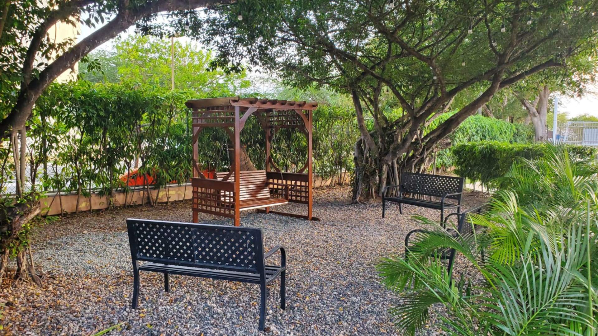 Wooden arbor with a swing and three metal benches in a graveled area surrounded by green trees and shrubs