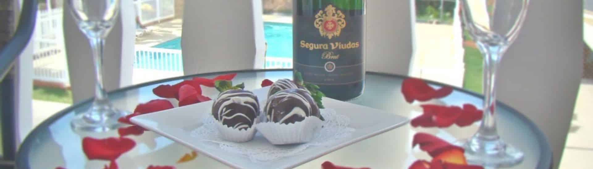 Close up view of chocolate covered strawberries on a square white plate, Champagne bottle, Champagne glasses, and red rose petals on a glass table